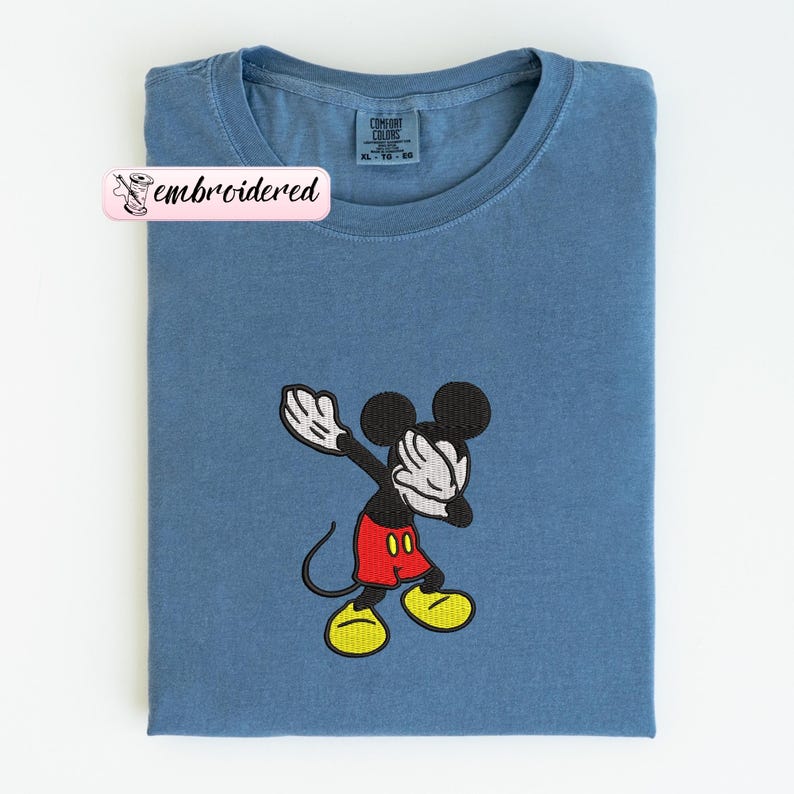 May include: A blue t-shirt with an embroidered Mickey Mouse design. The cartoon character is in a dabbing pose, with black ears, a black body, red shorts, yellow shoes, and white gloves. The shirt has a crew neck and a tag that says "Comfort Colors".