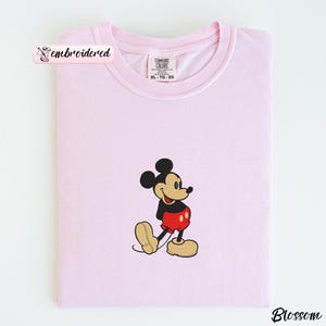 May include: A light pink t-shirt with a crew neck and short sleeves. The shirt features an embroidered image of Mickey Mouse in his classic outfit: black shorts, red pants, and yellow shoes. The shirt is folded neatly.