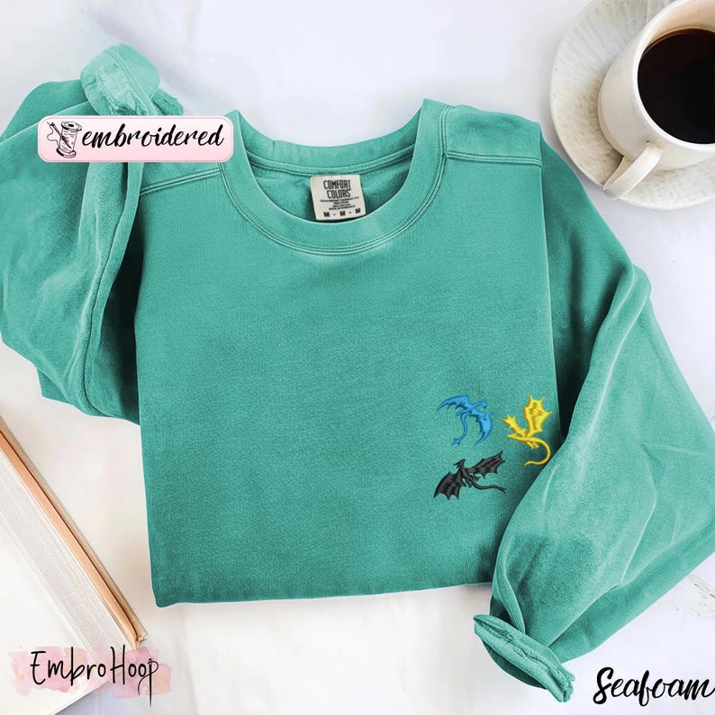 May include: Seafoam green sweatshirt with embroidered dragon designs in blue, yellow, and black. The sweatshirt has a crew neck and long sleeves. The word "embroidered" is in a pink banner on the top left. The word "EmbroHoop" is in a pink banner on the bottom left.