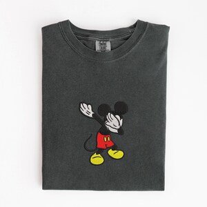 May include: Dark gray t-shirt with an embroidered image of Mickey Mouse dabbing. Mickey is wearing red shorts, yellow shoes, and white gloves. The shirt has a Comfort Colors tag.