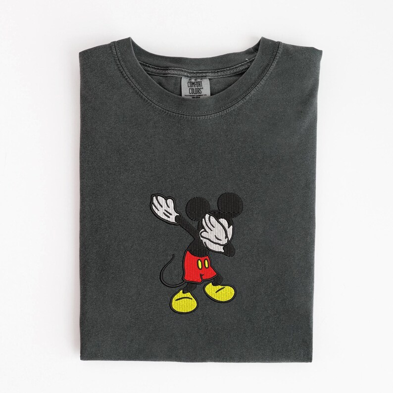 May include: Dark gray t-shirt with an embroidered image of Mickey Mouse dabbing. Mickey is wearing red shorts, yellow shoes, and white gloves. The shirt has a Comfort Colors tag.