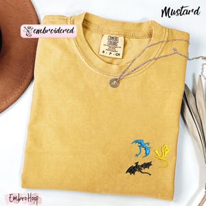 Pode incluir: Camiseta mostarda com drag&otilde;es bordados em azul, amarelo e preto. A camisa tem gola redonda e uma etiqueta pequena. Um colar de prata com um pingente est&aacute; drapeado sobre a camisa. A palavra "embroidered" est&aacute; em um adesivo rosa.