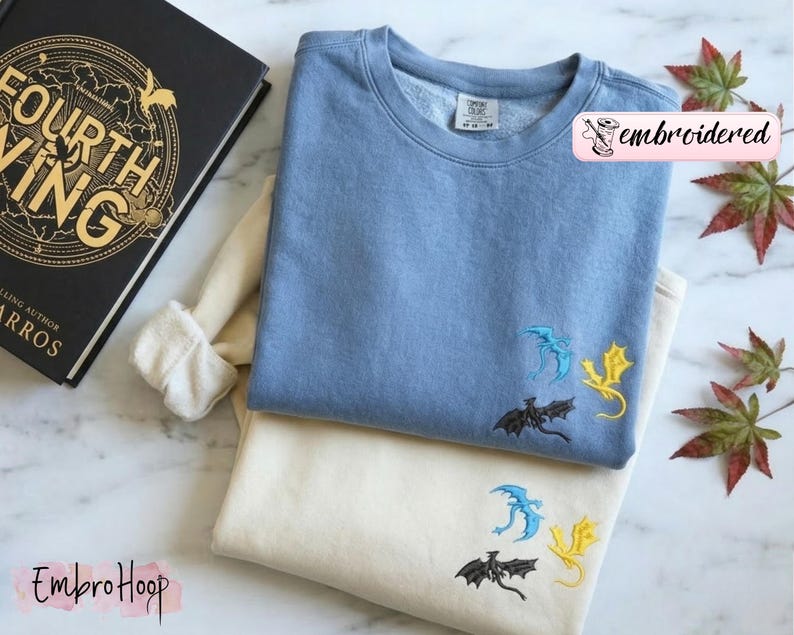 May include: Two sweatshirts, one light blue and one cream, are stacked. Each has three embroidered dragons in black, blue, and yellow. The light blue sweatshirt has the word "embroidered" in a pink banner. A book with gold lettering is visible.