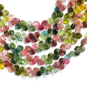 May include: Close-up of strands of colorful tourmaline gemstone beads. The beads are a mix of pink, green, yellow, and blue hues. Each bead is a small, faceted shape, and the strands are arranged in a slightly curved pattern against a white background.