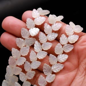 May include: A collection of small, translucent white leaf-shaped beads. Each bead has detailed veining, and the leaves are clustered in groups. The beads are held in a hand against a black background, showcasing their delicate design.
