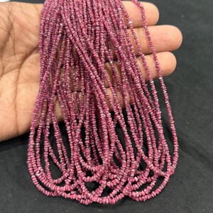 May include: A long strand of small, raw, deep pink garnet beads. The beads are irregular in shape and texture, featuring a faceted, natural appearance. The strand is composed of numerous tiny, unpolished stones strung together in a continuous loop.
