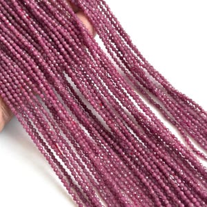 Peut inclure: Plusieurs longs brins de petites perles en pierre précieuse facettées de couleur magenta profond. Chaque perle individuelle est ronde et présente une surface texturée et réfléchissante qui capte la lumière, disposée en lignes parallèles.