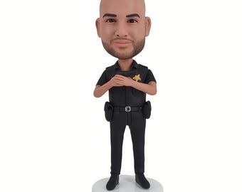 May include: A bobblehead figurine of a police officer in a black uniform, with a gold star badge. The figurine has a bald head and is standing on a white base with the name "OFFICER OLIVIA" printed on it.