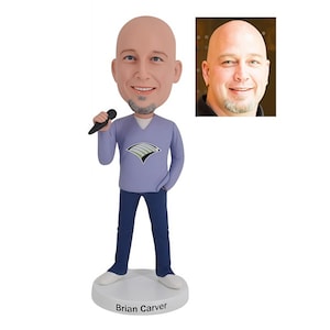 May include: A custom bobblehead figurine of a man holding a black microphone. The figure wears a light purple sweater, dark blue trousers, and white shoes. The base is white with the name "Brian Carver" printed on it. A smaller photo of the man is in the upper right corner.