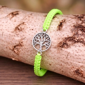 Può includere: Un braccialetto in macramè verde neon con un ciondolo albero della vita color argento. Il braccialetto è esposto su un pezzo di legno, mettendo in risalto il colore verde brillante del cordoncino intrecciato e il design dettagliato del ciondolo.