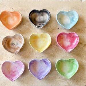 Könnte beinhalten: Neun herzförmige Schmuckschalen in verschiedenen Farben, darunter Orange, Schwarz, Blau, Braun, Gelb, Rosa, Lila und Grün. Jede Schale hat ein marmoriertes Aussehen und steht auf einer hellen, strukturierten Oberfläche.