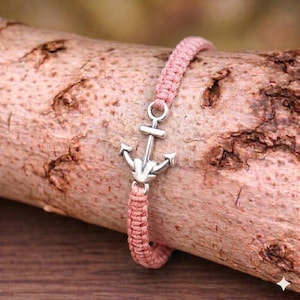 Pode incluir: Uma pulseira de macramé rosa com um berloque de âncora prateada. A pulseira é feita de cordão rosa tecido e apresenta um pingente de âncora prateada detalhado. A pulseira é exibida em um pedaço de madeira.