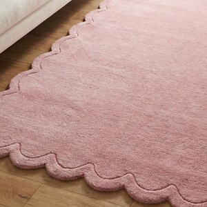 May include: A soft, pink rug with a wavy, scalloped edge. The rug is a solid colour and has a fluffy texture. The rug is placed on a wooden floor.