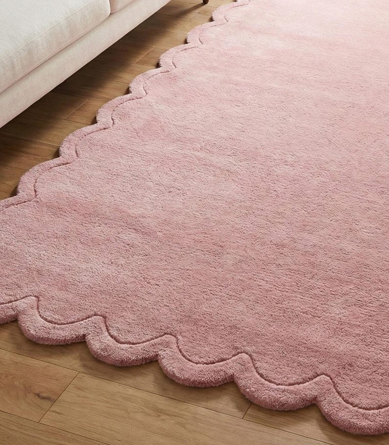 May include: A soft, pink rug with a wavy, scalloped edge. The rug is a solid colour and has a fluffy texture. The rug is placed on a wooden floor.