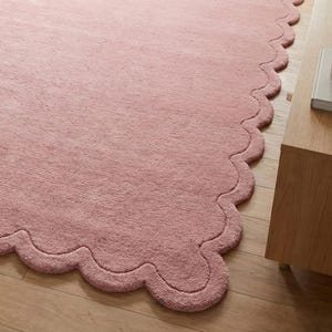 May include: A pink rug with a wavy, scalloped edge. The rug has a soft, plush texture and is placed on a wooden floor. A wooden side table is visible in the corner. The rug is a decorative home accent.