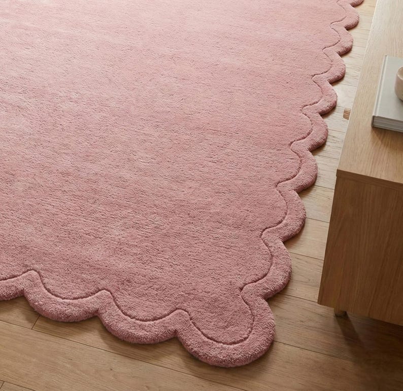 May include: A pink rug with a wavy, scalloped edge. The rug has a soft, plush texture and is placed on a wooden floor. A wooden side table is visible in the corner. The rug is a decorative home accent.