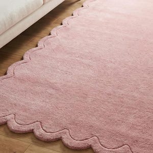 May include: A pale pink rug with a distinctive scalloped edge. The rug is made of a soft, fluffy material and is placed on a wooden floor. The rug's design adds a touch of elegance and softness to the room.
