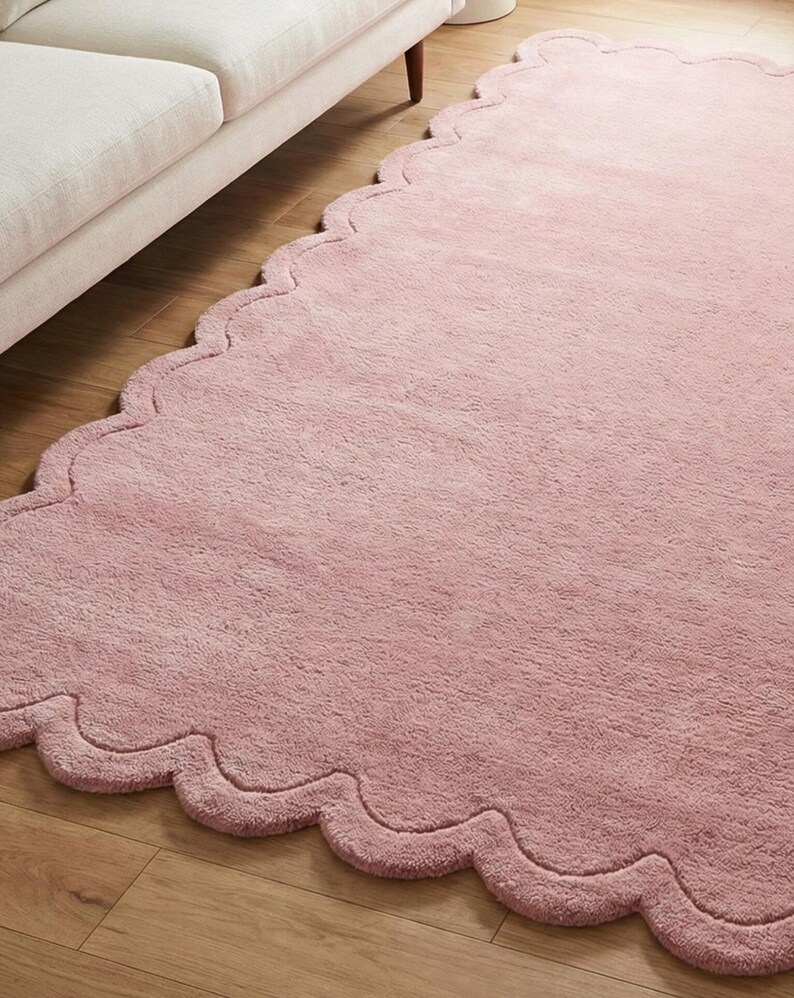 May include: A pale pink rug with a distinctive scalloped edge. The rug is made of a soft, fluffy material and is placed on a wooden floor. The rug's design adds a touch of elegance and softness to the room.