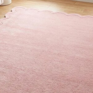 May include: A soft, pink, scalloped-edge rug. The rug has a plush texture and is placed on a light-coloured wooden floor. The rug's shape is a large rectangle with a wavy, decorative border. The rug is near a white sofa.