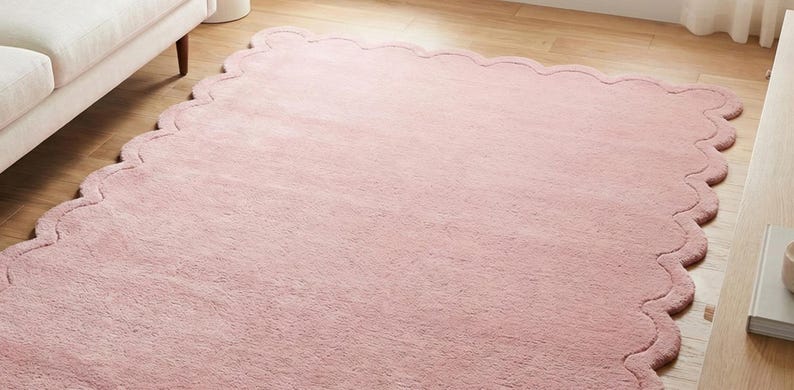 May include: A soft, pink, scalloped-edge rug. The rug has a plush texture and is placed on a light-coloured wooden floor. The rug's shape is a large rectangle with a wavy, decorative border. The rug is near a white sofa.