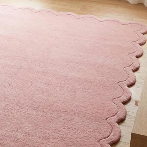 May include: A large, soft, pink rug with a scalloped edge. The rug is made of a plush material and is placed on a light-coloured wooden floor. The rug's design adds a touch of elegance to the room.