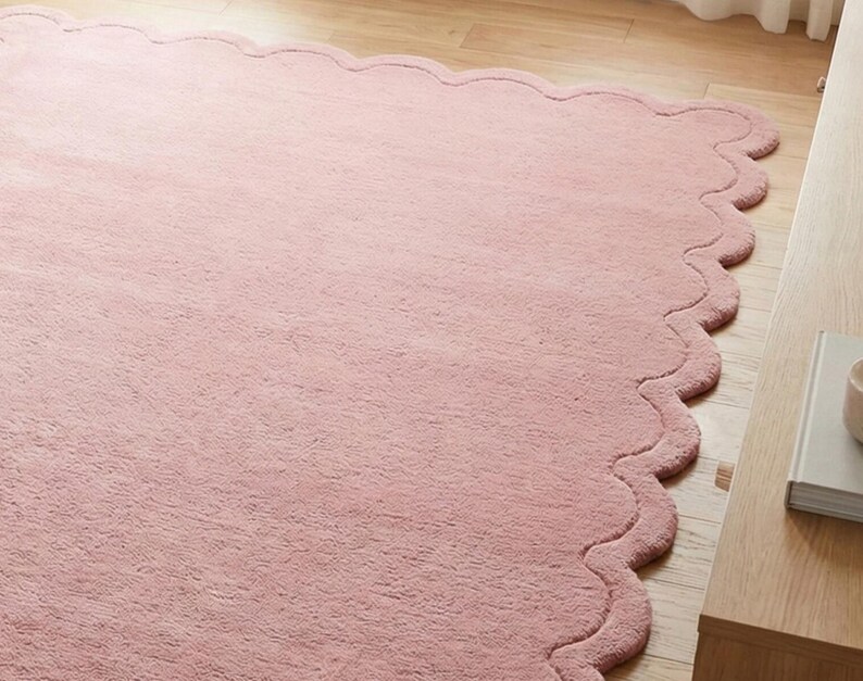 May include: A large, soft, pink rug with a scalloped edge. The rug is made of a plush material and is placed on a light-coloured wooden floor. The rug's design adds a touch of elegance to the room.