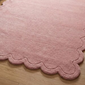 May include: A large, soft, pink rug with a scalloped edge. The rug is made of a plush material and is placed on a wooden floor. The rug's shape is irregular, with a wavy border.