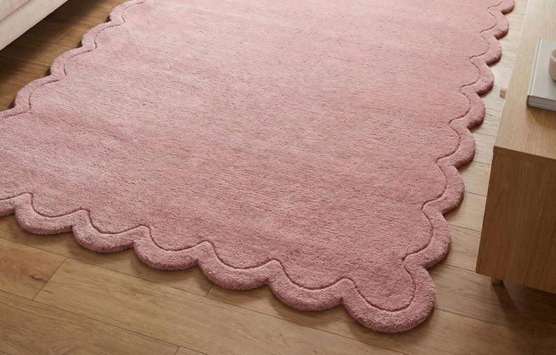 May include: A large, soft, pink rug with a scalloped edge. The rug is made of a plush material and is placed on a wooden floor. The rug's shape is irregular, with a wavy border.