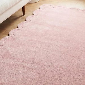 May include: A pale pink rug with a scalloped border. The rug is on a wooden floor, next to a white sofa. The rug has a soft, fluffy texture, adding a touch of elegance to the room. The rug's dimensions are in centimeters.