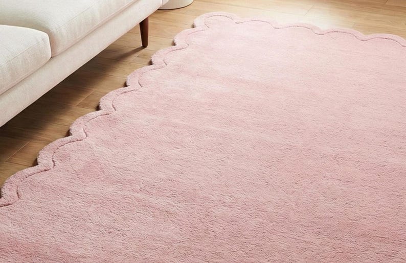 May include: A pale pink rug with a scalloped border. The rug is on a wooden floor, next to a white sofa. The rug has a soft, fluffy texture, adding a touch of elegance to the room. The rug's dimensions are in centimeters.