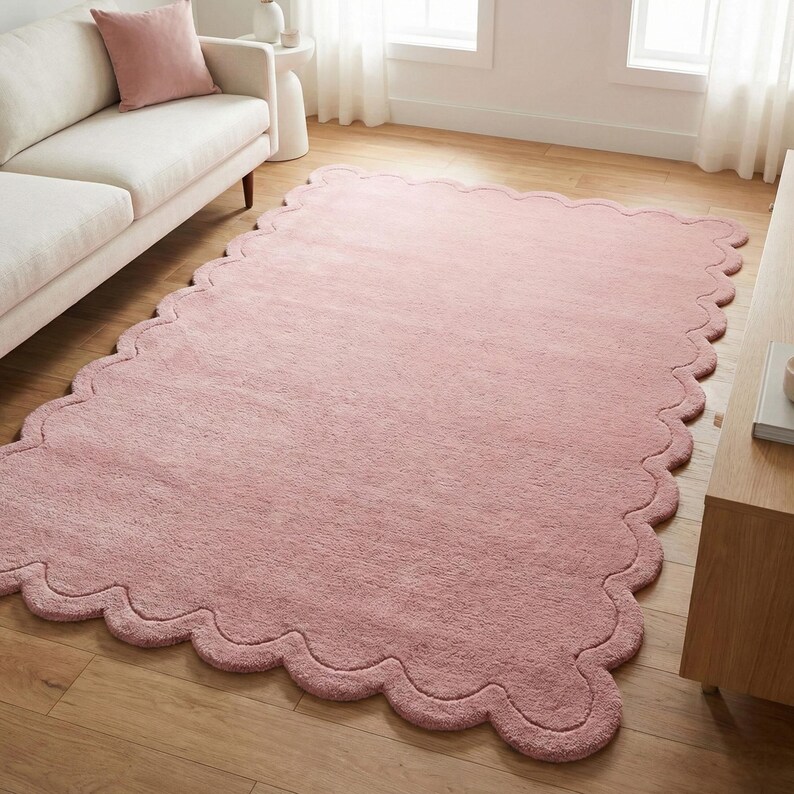 May include: A large, rectangular, dusky pink rug with a scalloped edge. The rug is made of a soft, plush material and is placed on a wooden floor. The rug is in a living room setting.