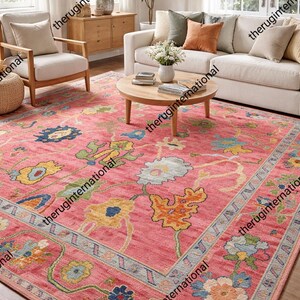 Pode incluir: Um tapete rosa retangular grande com um padrão floral em tons de azul, amarelo, laranja e verde. O tapete está colocado numa sala de estar com um sofá branco, cadeiras e uma mesa de centro de madeira.