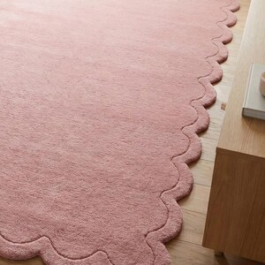May include: A large, soft, pink rug with a scalloped edge. The rug is made of a plush material and is placed on a wooden floor. The rug's design adds a touch of elegance to the room.