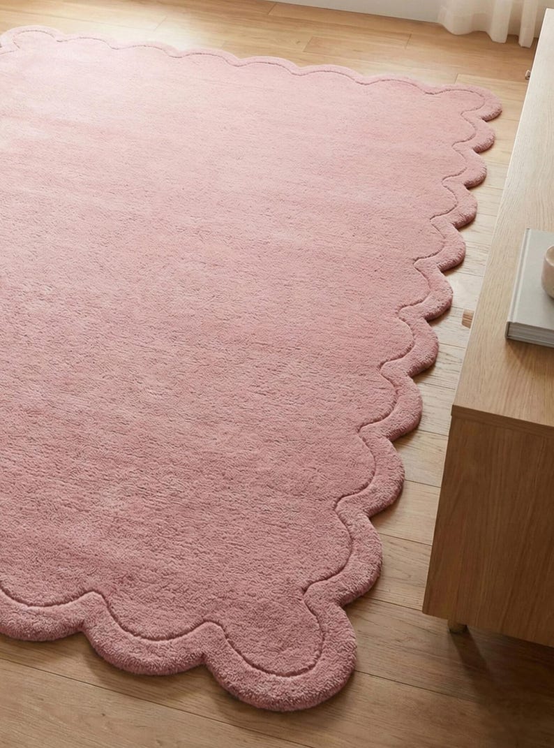 May include: A large, soft, pink rug with a scalloped edge. The rug is made of a plush material and is placed on a wooden floor. The rug's design adds a touch of elegance to the room.