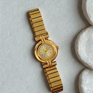 May include: A gold-toned wristwatch with a round face and a stretch band. The watch face features Roman numerals and the brand name "Lutz". The watch is set against a white background.