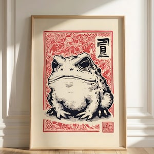May include: Framed art print featuring a detailed black and white illustration of a toad. The artwork is set against a cream background with a red decorative border and Japanese characters. The print is encased in a light wood frame.
