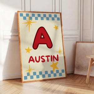 May include: A framed art print with the name "Austin" in red, a large red letter "A", and yellow star accents. The print has a cream background with a blue and white checkered border. The frame is a warm brown colour.