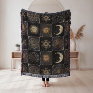 May include: Black throw blanket with a repeating pattern of celestial symbols, including suns, moons, and geometric shapes. The blanket has a fringed edge and a dark color palette with gold accents. The blanket is held up by a person.