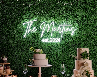 May include: A white neon sign featuring the cursive text The Martins and the printed text est. 2024. The sign is mounted on a dense green artificial hedge wall. The neon tubing emits a bright white glow, creating a clear contrast against the foliage.