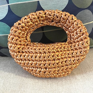 Puede incluir: Bolso de mano artesanal de color dorado metálico con textura tejida y asa circular. El bolso tiene forma ovalada y redondeada. Se muestra sobre una superficie gris claro, con un fondo estampado azul y verde.