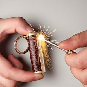 Op de afbeelding: Een bruine en gouden aansteker met een decoratief patroon wordt aangestoken. De aansteker heeft een sleutelringbevestiging en het woord "HONEST" is zichtbaar. Heldere vonken komen uit de aansteker als deze wordt aangestoken.