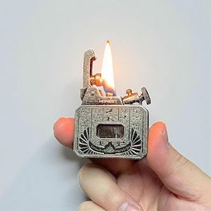 Może przedstawiać: Zapalniczka w stylu vintage z motywem egipskim, z misternymi grawerunkami i płomieniem. Zapalniczka jest w kolorze srebrnym, z prostokątnym korpusem i klapką na zawiasach. Widoczny jest napis "HONEST".