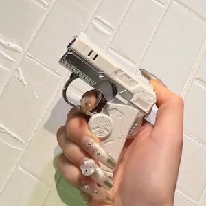 Op de afbeelding: Een wit en zilverkleurig aansteker in de vorm van een pistool. De aansteker wordt vastgehouden in een hand met lange, versierde nagels. De aansteker heeft een trekker, een loop en andere details die een echt vuurwapen nabootsen.