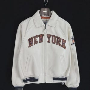 May include: Cream-colored leather jacket with the text "NEW YORK" in brown and orange lettering across the chest. The jacket has a zip-up front, a collar, and striped cuffs and hem. A small patch with a flag design is on the sleeve.