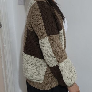 May include: A patchwork crochet cardigan in shades of brown and cream. The open-front sweater features a geometric design with rectangular panels of varying colors. The sleeves are long and the overall style is casual and handmade.