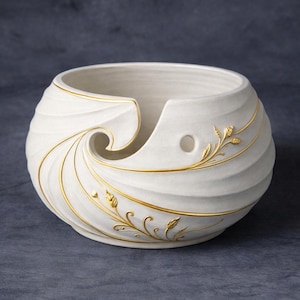 May include: A white ceramic yarn bowl with a swirling design and gold accents. The bowl features a spiral opening and a small hole for the yarn. Gold leaf and vine details adorn the exterior, adding a touch of elegance to this craft accessory.