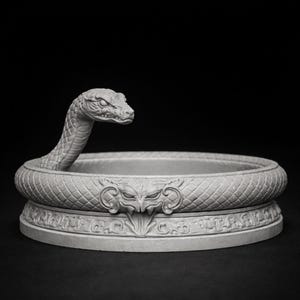 May include: A gray, detailed sculpture of a snake coiled around a circular base. The snake's head is raised, and the base features a decorative design with a central face. The sculpture is made of a light-colored material and is set against a dark background.