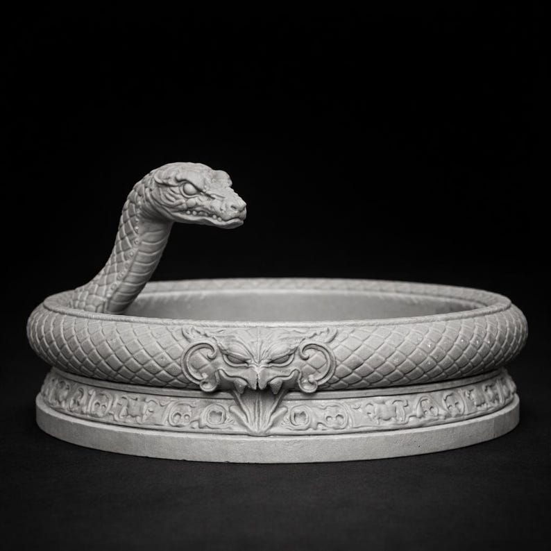 May include: A gray, detailed sculpture of a snake coiled around a circular base. The snake's head is raised, and the base features a decorative design with a central face. The sculpture is made of a light-colored material and is set against a dark background.