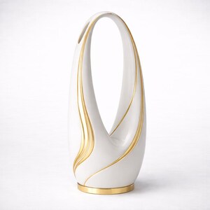 May include: A white ceramic vase with a gold base and gold detailing. The vase has a distinctive, elongated form with a handle-like top. The gold accents cascade down the sides, enhancing the vase's elegant design. A decorative item.