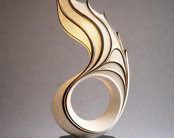 May include: A sculptural table lamp featuring a curved, flame-like form with a white, textured, perforated surface and black edges. The structure rises from a circular black base, creating an open, circular void in the center.